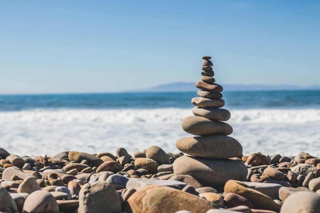 A stack of smooth stones balanced on a rocky beach with the ocean and a distant island in the background under a clear blue sky.